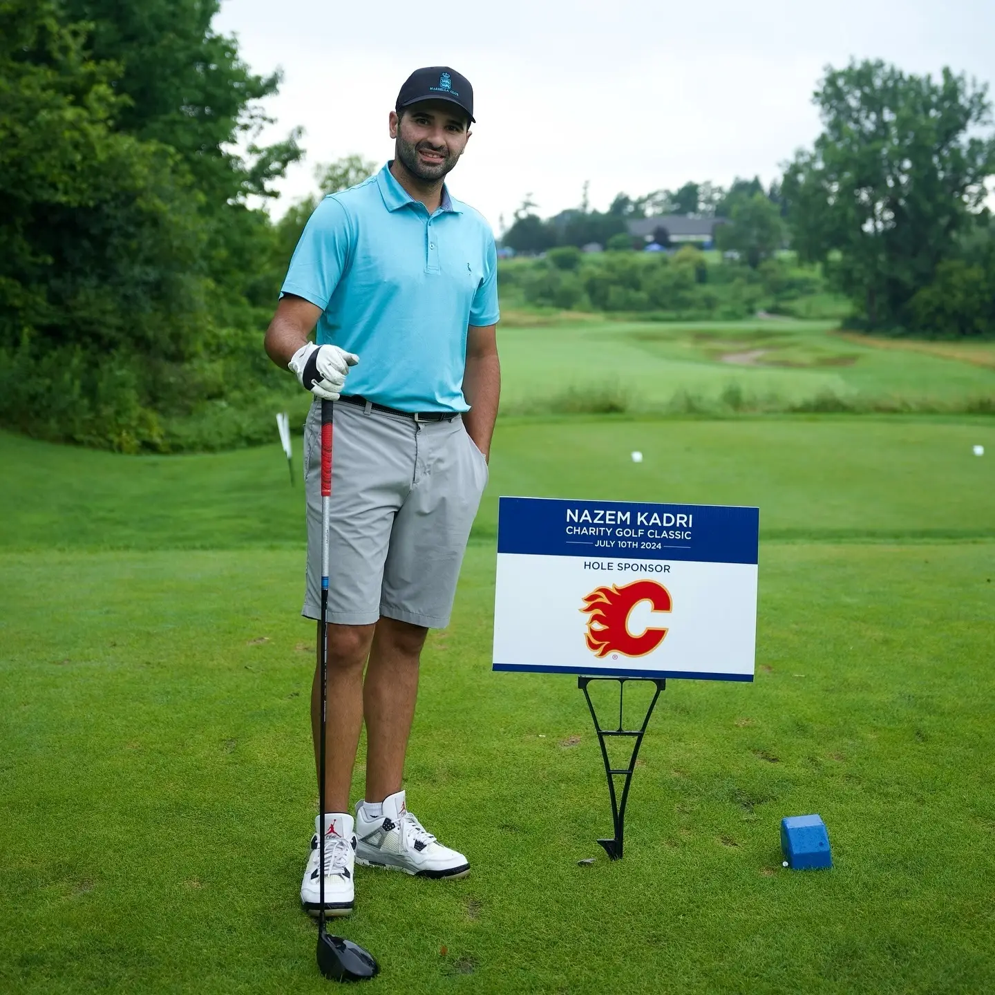 Male golfer in a light blue polo and dark cap stands with a putter on a green golf course beside a sponsor sign for Nazem Kadri Charity Golf Classic.